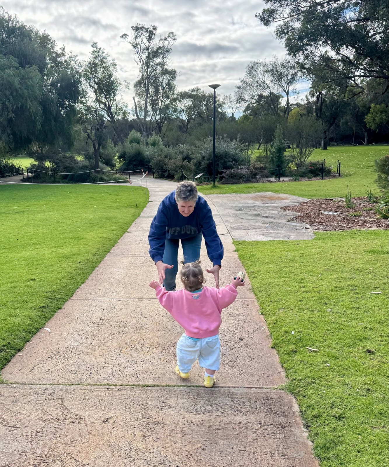 A grandmother crouching with arms out as a toddler walks towards her along a park path